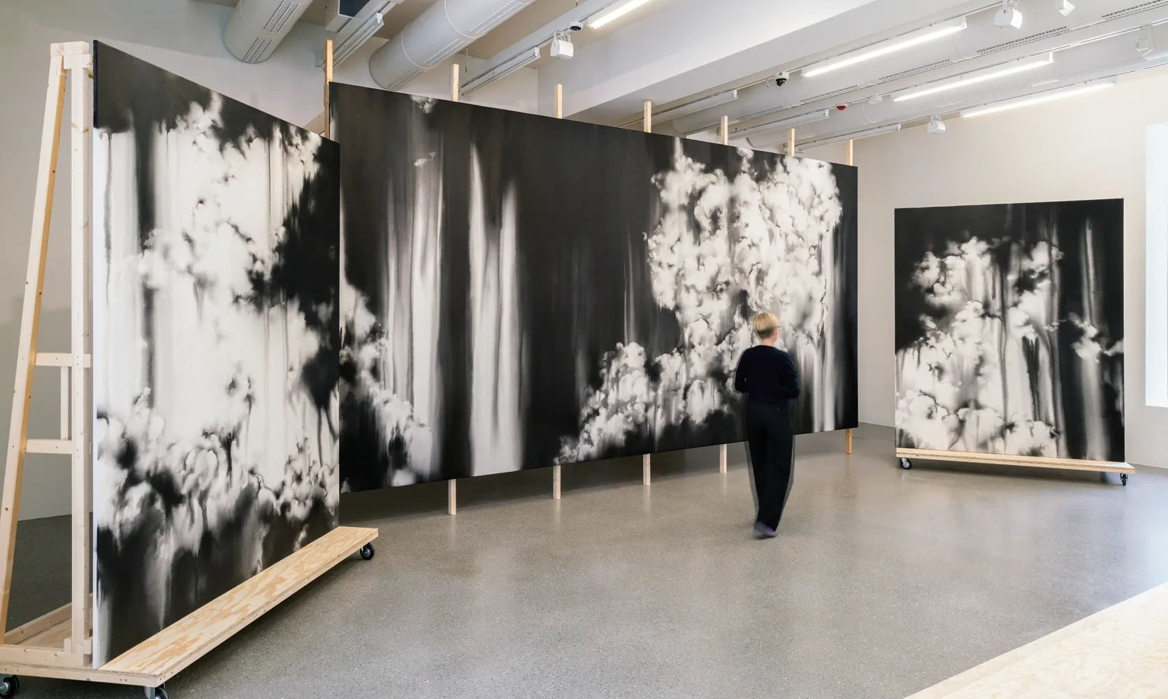 Lucia Tallová, PalaisPopulaire, Berlin: large black and white cloud paintings on wheeled wooden frames, a visitor standing before them.