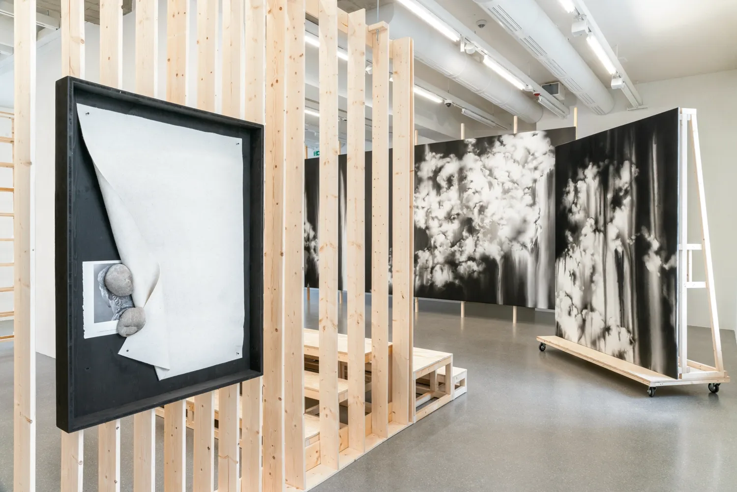 Lucia Tallová, PalaisPopulaire, Berlin: black-framed work with curling cream paper and a stone before a slatted wall and cloud paintings.