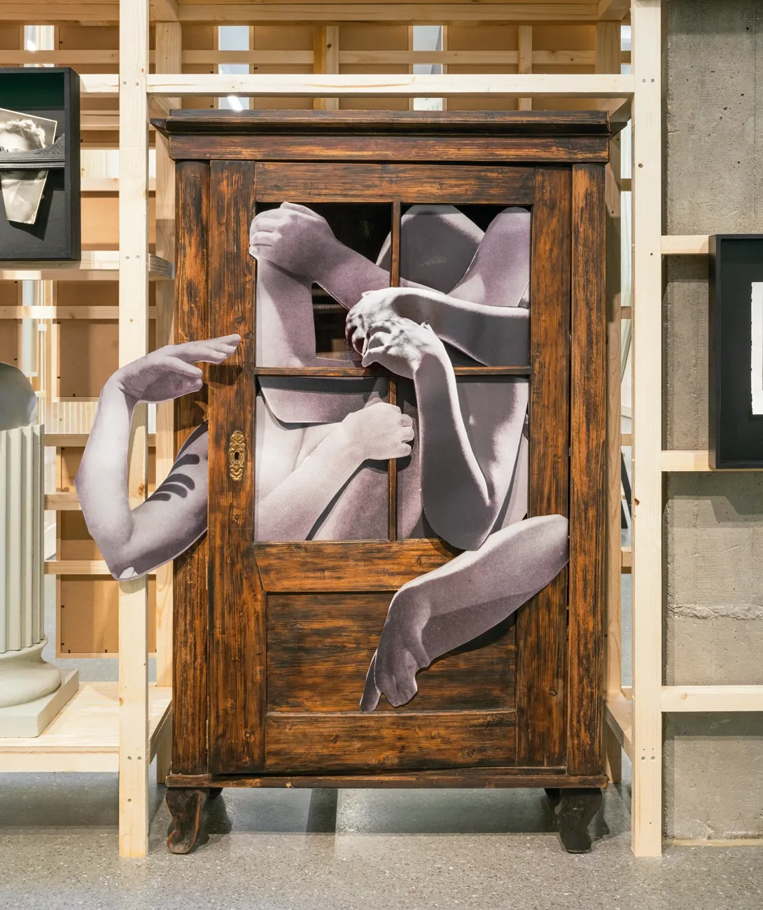 Lucia Tallová, PalaisPopulaire, Berlin: dark wooden cabinet with purple-toned printed arms and legs emerging from its glazed door.