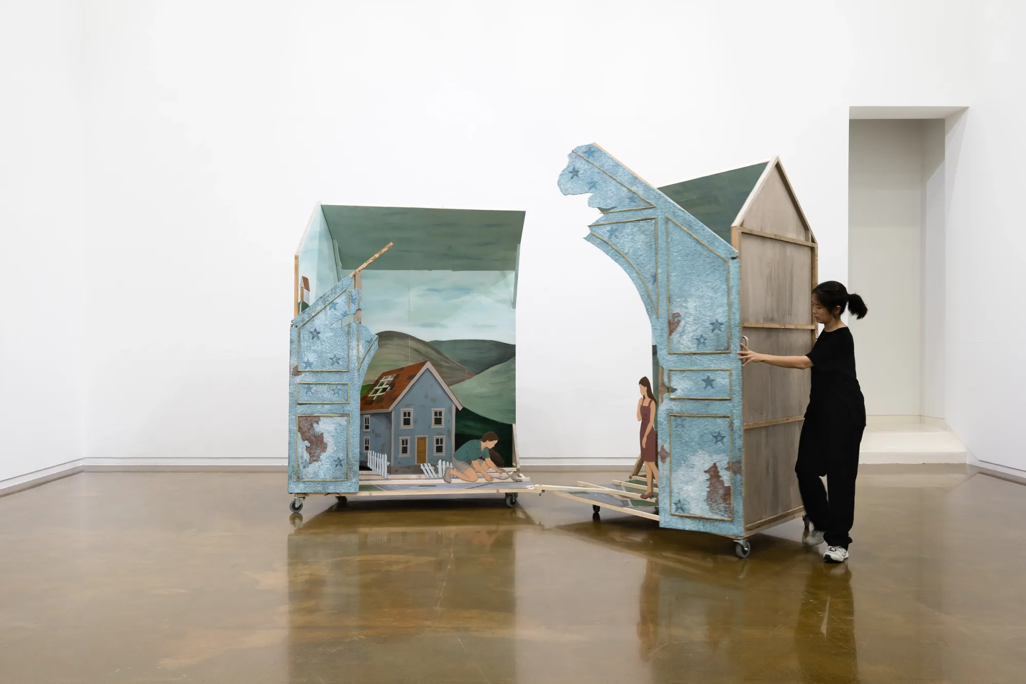 ALT: Seongeun Lee, Cheongju Art Studio, Cheongju: a person in black pushes one half of a split house on casters, painted figures in the other half.
