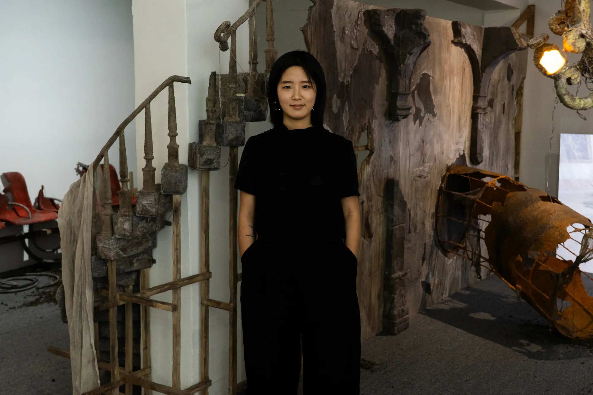  Seongeun Lee, artist portrait: catapult contemporary vienna, a woman artist female in black clothes stands in front of a weathered wooden wall and a rusted metal sculpture.