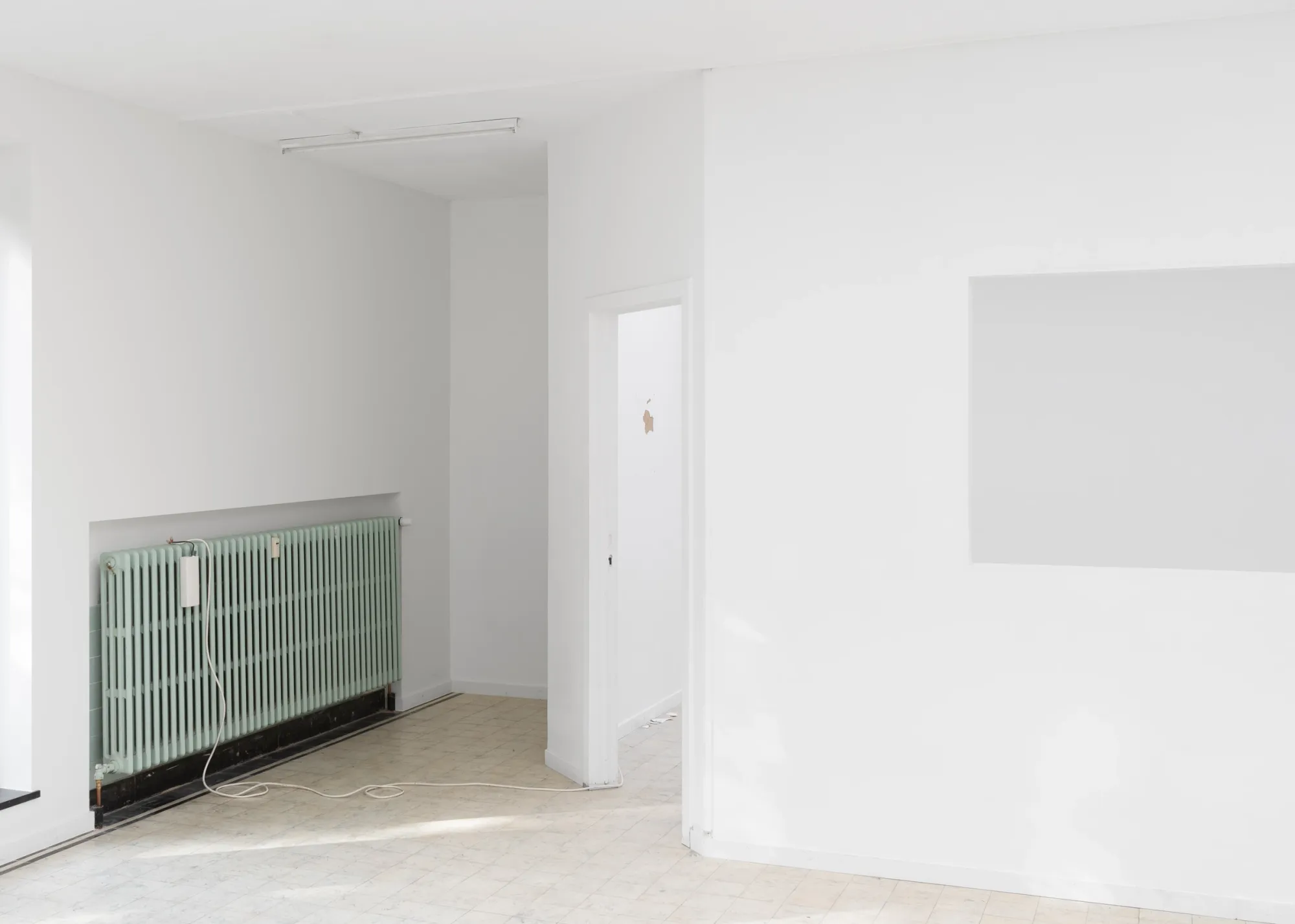 Exhibition review of Simon Bultynck: A. Aubry, A. Colin, P. Lippens, A. Oosterlinck, C. Wynants at CONVENT Space for Contemporary Art, Ghent, 2026. White room with a pale green radiator, a white box and cable running across the tiled floor, beside a doorway and a wall opening.