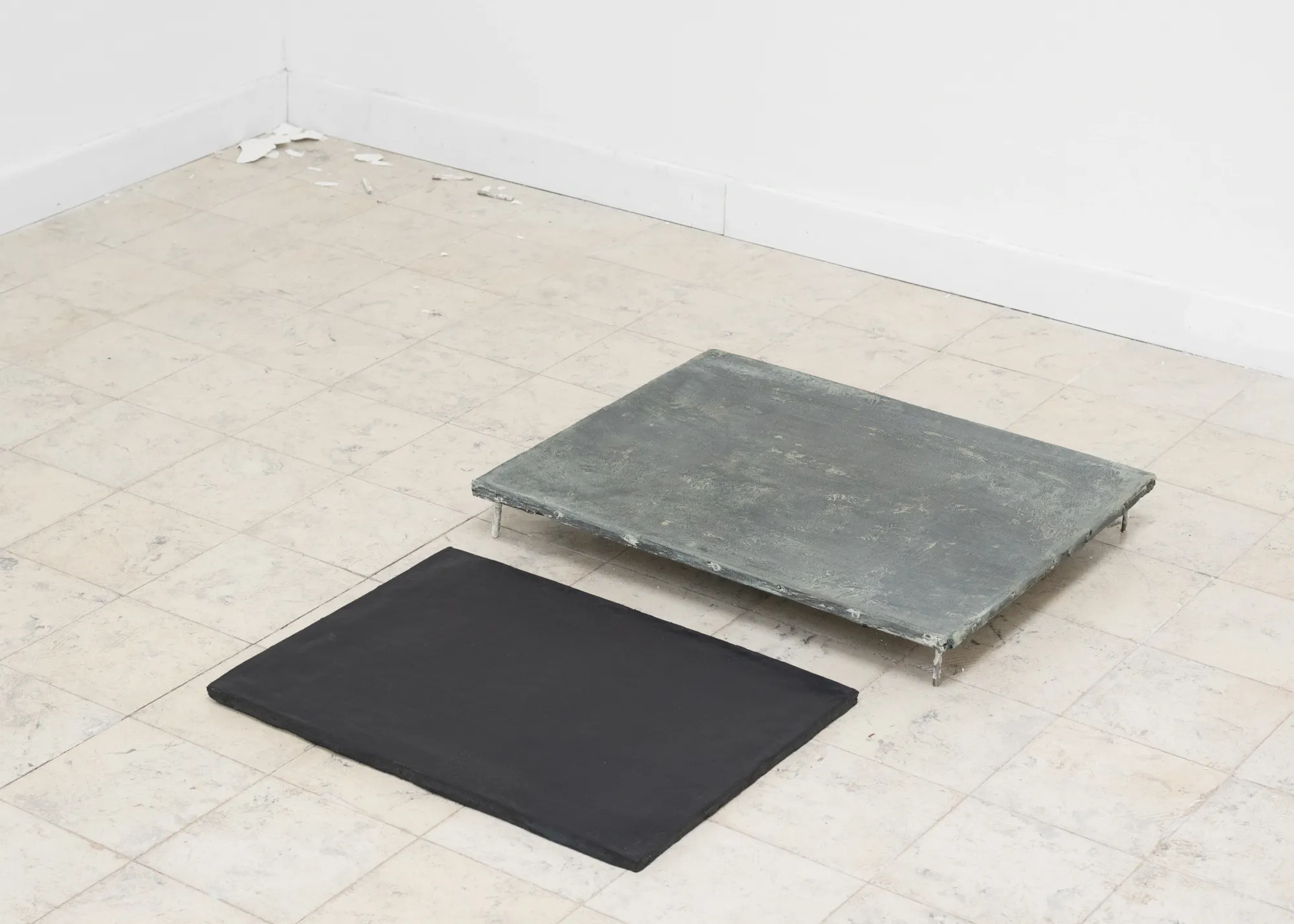 Exhibition review of Simon Bultynck: A. Aubry, A. Colin, P. Lippens, A. Oosterlinck, C. Wynants at CONVENT Space for Contemporary Art, Ghent, 2026. Two flat works on a tiled floor, one matte black and one grey-green on short metal legs, near a white corner.