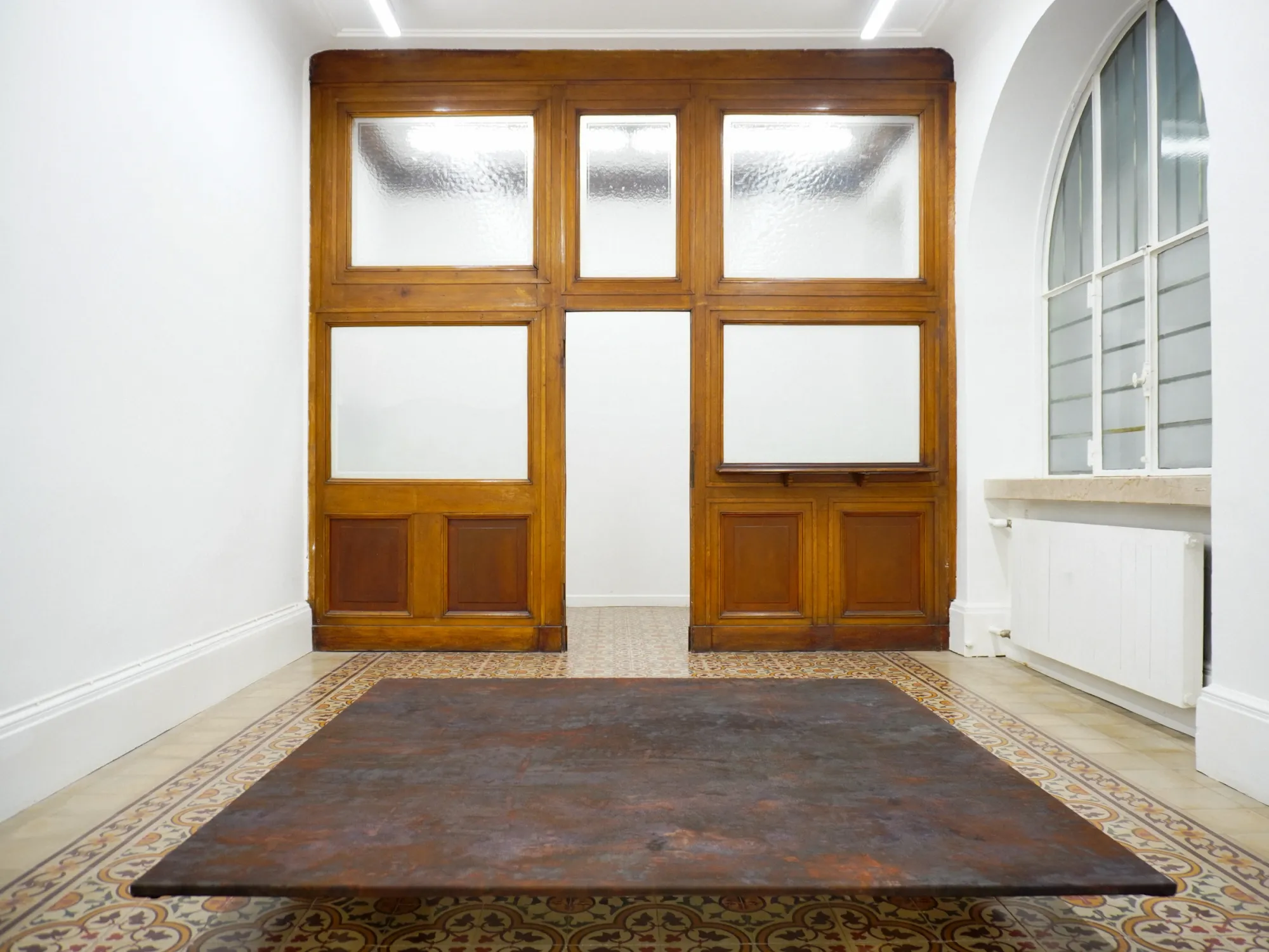 Taylah Hasaballah, INDICATRIX at The Address, Brescia, 2026. Low rust-brown panel before a wooden door with frosted glass.