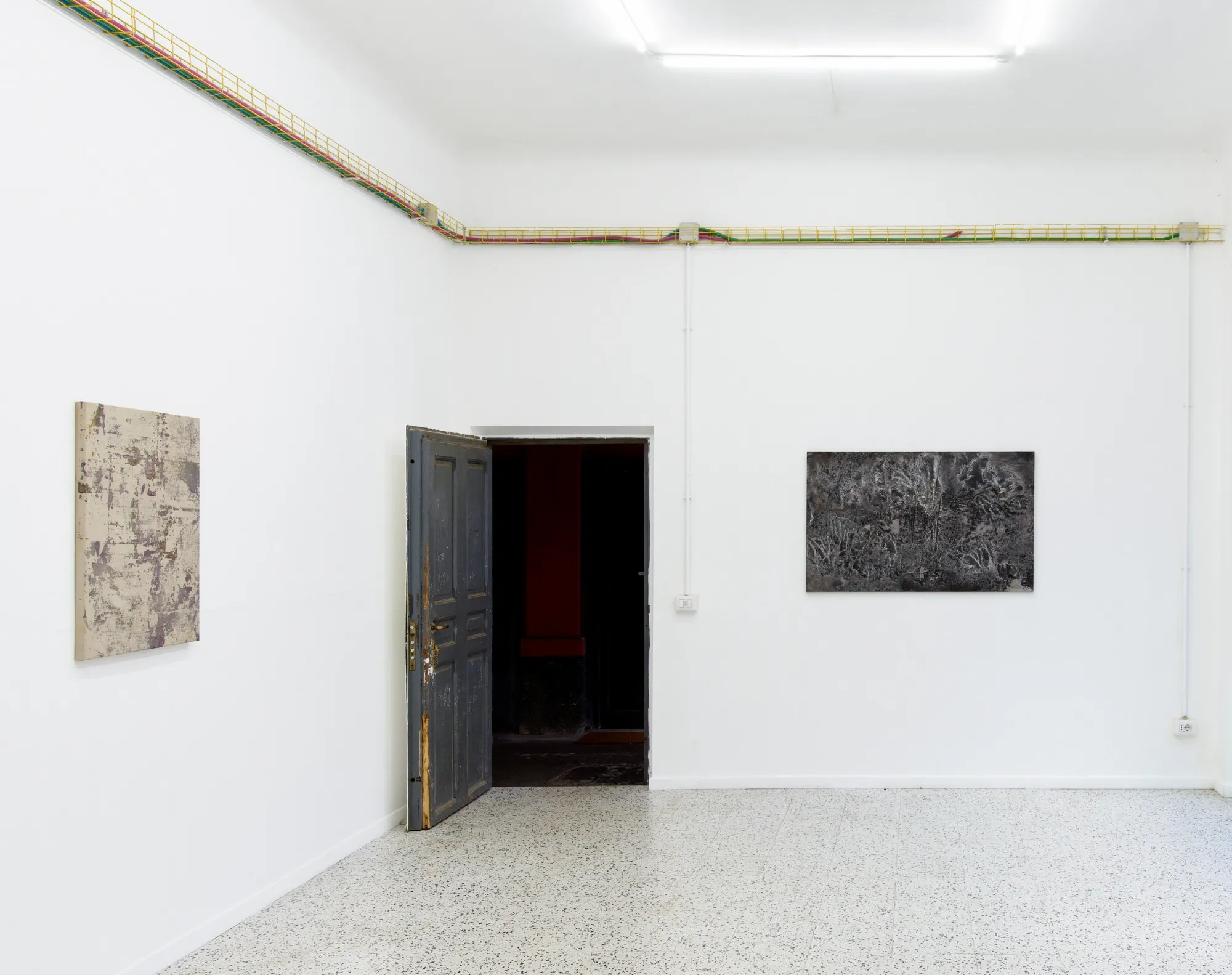 Taylah Hasaballah, INDICATRIX at The Address, Milan, 2026. Beige panel and dark textured painting on a white wall around an open door.