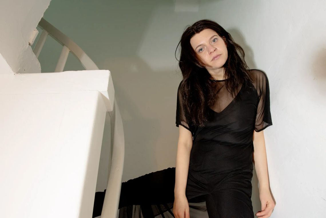 Portrait of Nadine Karl standing beside a sculptural installation in a gallery space, wearing a black top, shown in relation to her work.