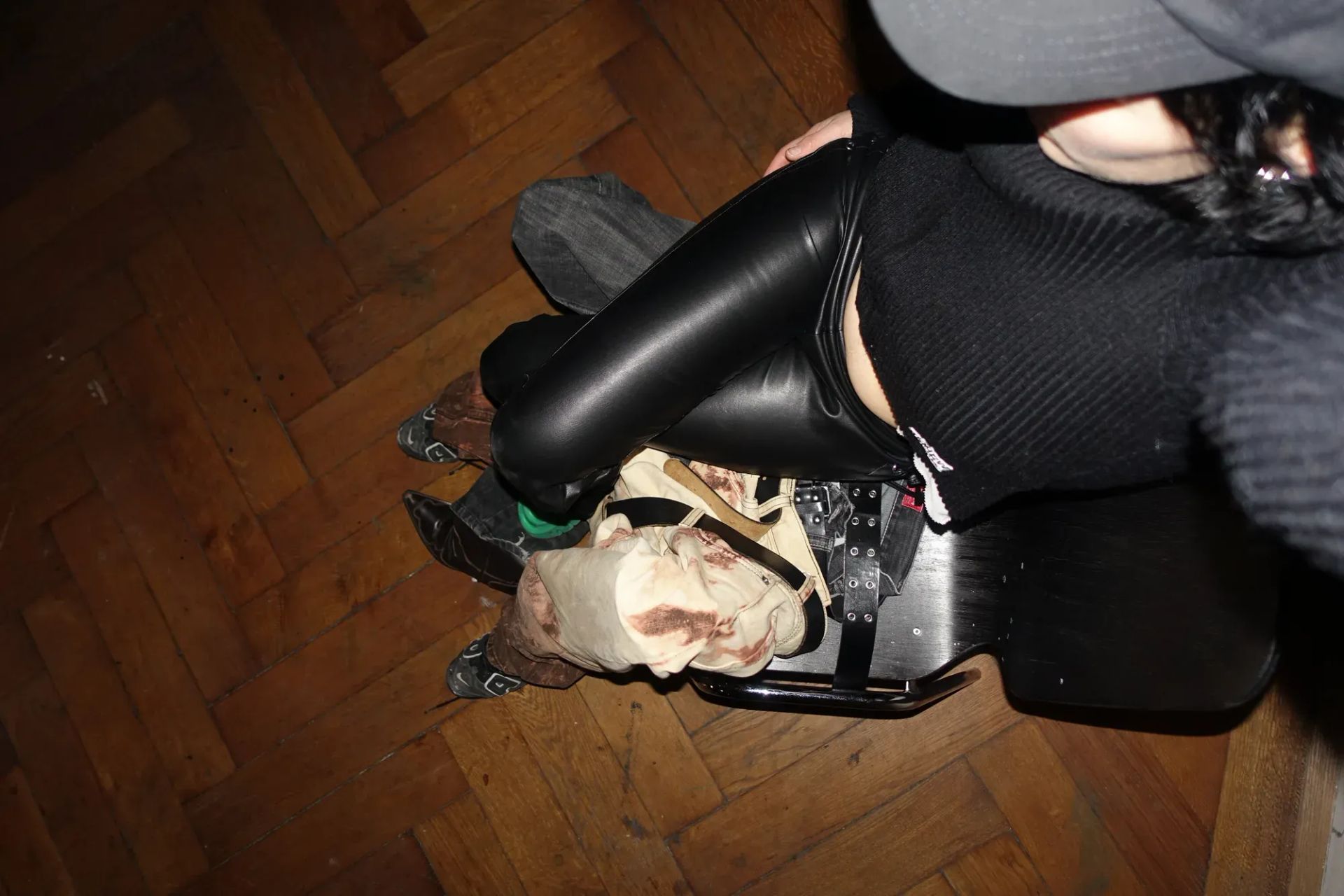 Ju Aichinger aus der Vogelperspektive; Lederhose, Stiefel, auf dem Schoß ein skulpturales Kostümteil mit Riemen und Flecken, Parkettboden – Feature Munchies Art Club. – Wien, AT.