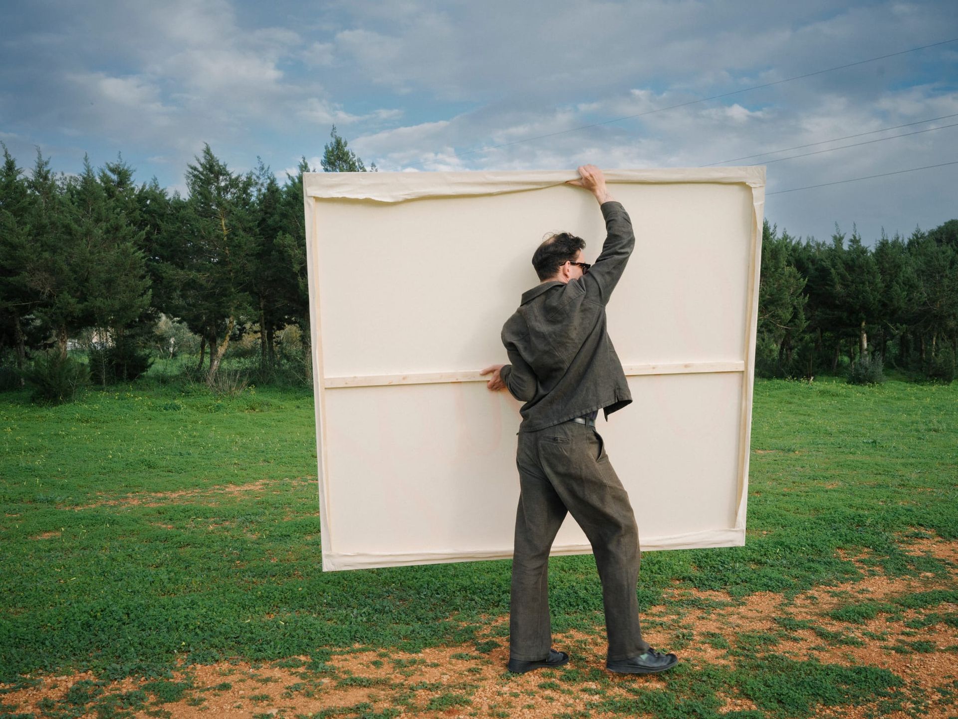 Tom Król standing outdoors holding the back of a large blank canvas against a landscape of trees and sky.