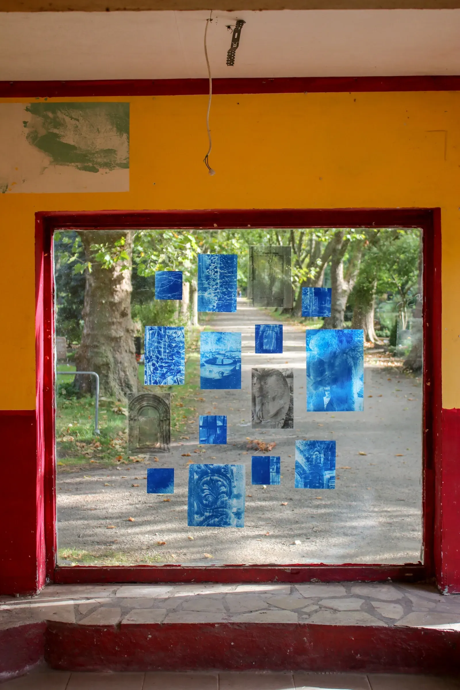 Aubrey Theobald and Kristina Popov, Phantom Pförtnerhaus Chapter 3, Berlin, 2026. Small blue and grey photographic prints on a window pane overlooking a cemetery path.