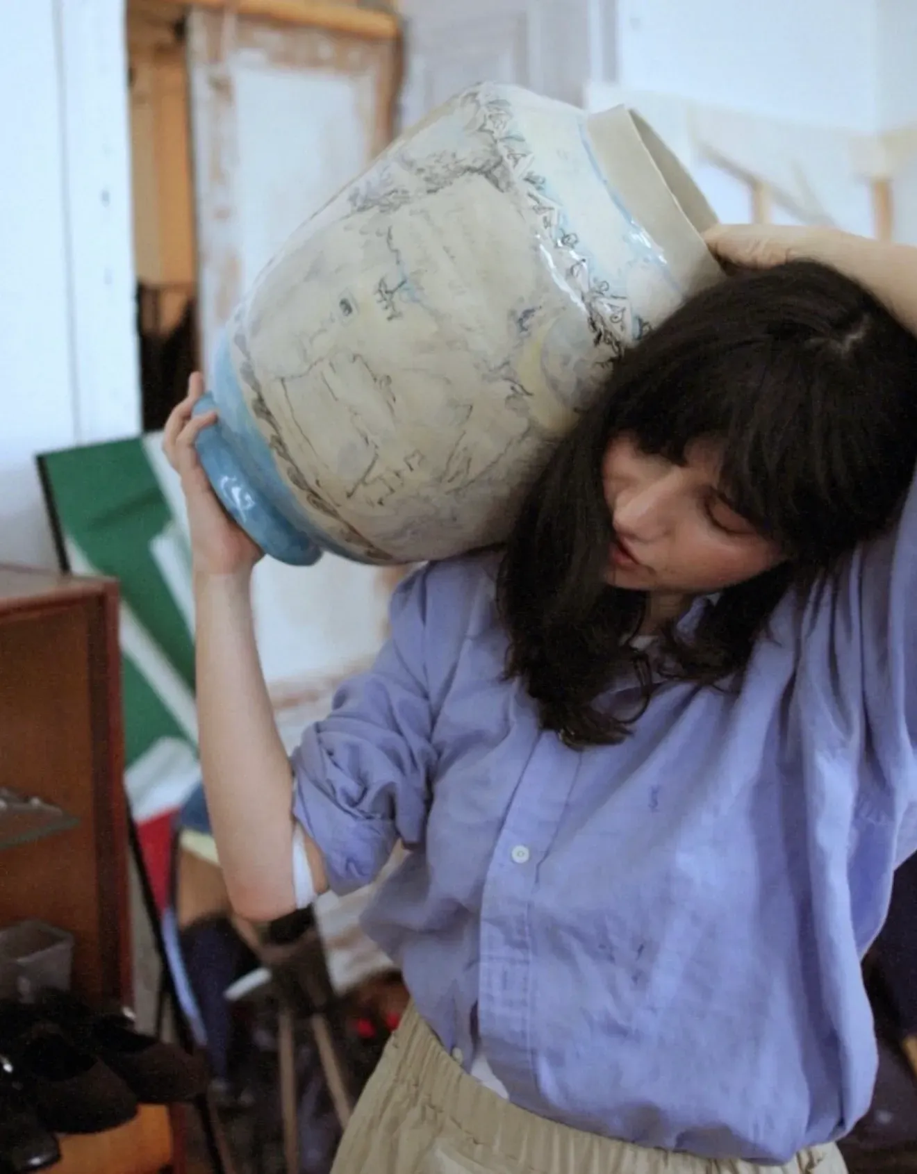 Interview with the artist Christina Arark: artist in a blue shirt carrying a ceramic vase on her shoulder in the studio.