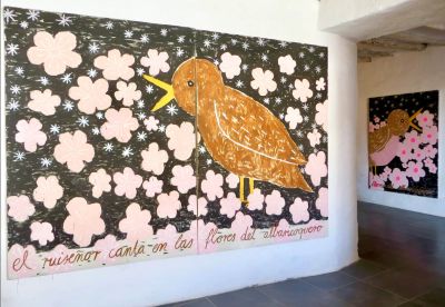 Studio view with two large Gabrielle Grässle canvases: brown bird amid pink blossoms with a handwritten Spanish line; second related painting visible in the adjacent white-walled room.