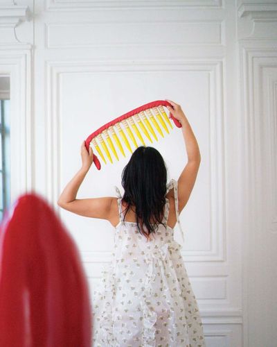 Eri Maeda lifting her large red and yellow ceramic Monster Comb sculpture in a white Paris room, wearing a floral Cecilie Bahnsen dress. Contemporary ceramic art and self presentation.