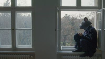 Exhibition review of Marwa Arsanios: Who is Afraid of Ideology? at TAXISPALAIS Kunsthalle Tirol, Innsbruck, 2026. Figure in a grey mouse mask sitting on a windowsill overlooking a cemetery.