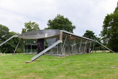 Reparaturgesellschaft, Wewerka Pavilion, Münster, 2026, glass and steel pavilion on grass with pink graffiti on black door, visitors nearby