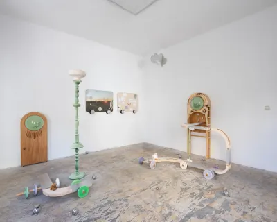 Exhibition review of Sebastian Scholz: ludo at global international, Vienna, 2026. Green standing lamp, wooden porthole wardrobe and wheeled waggon sculptures