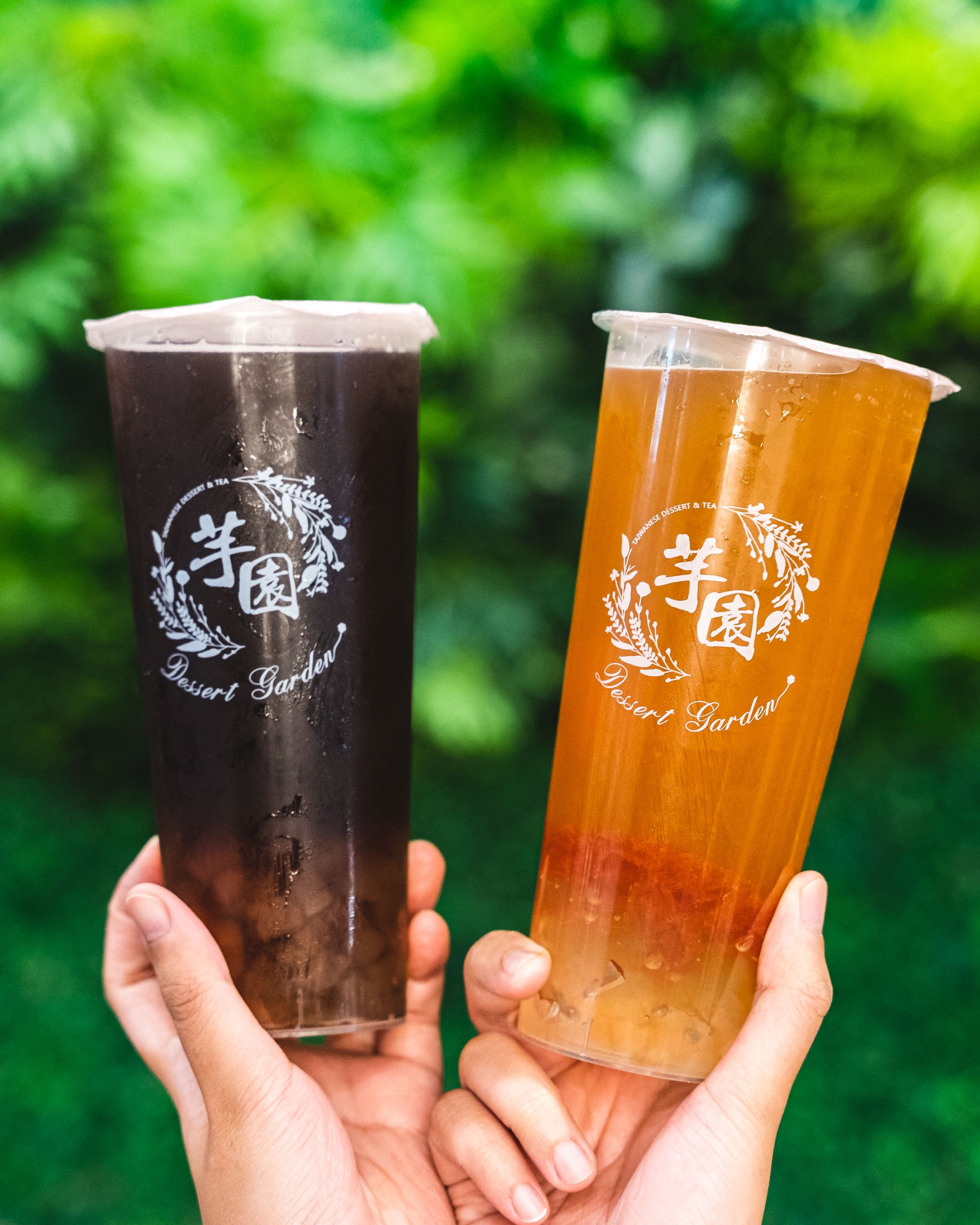 Hands holding two tea drinks in a sealed plastic cup in-front of a blurred green background