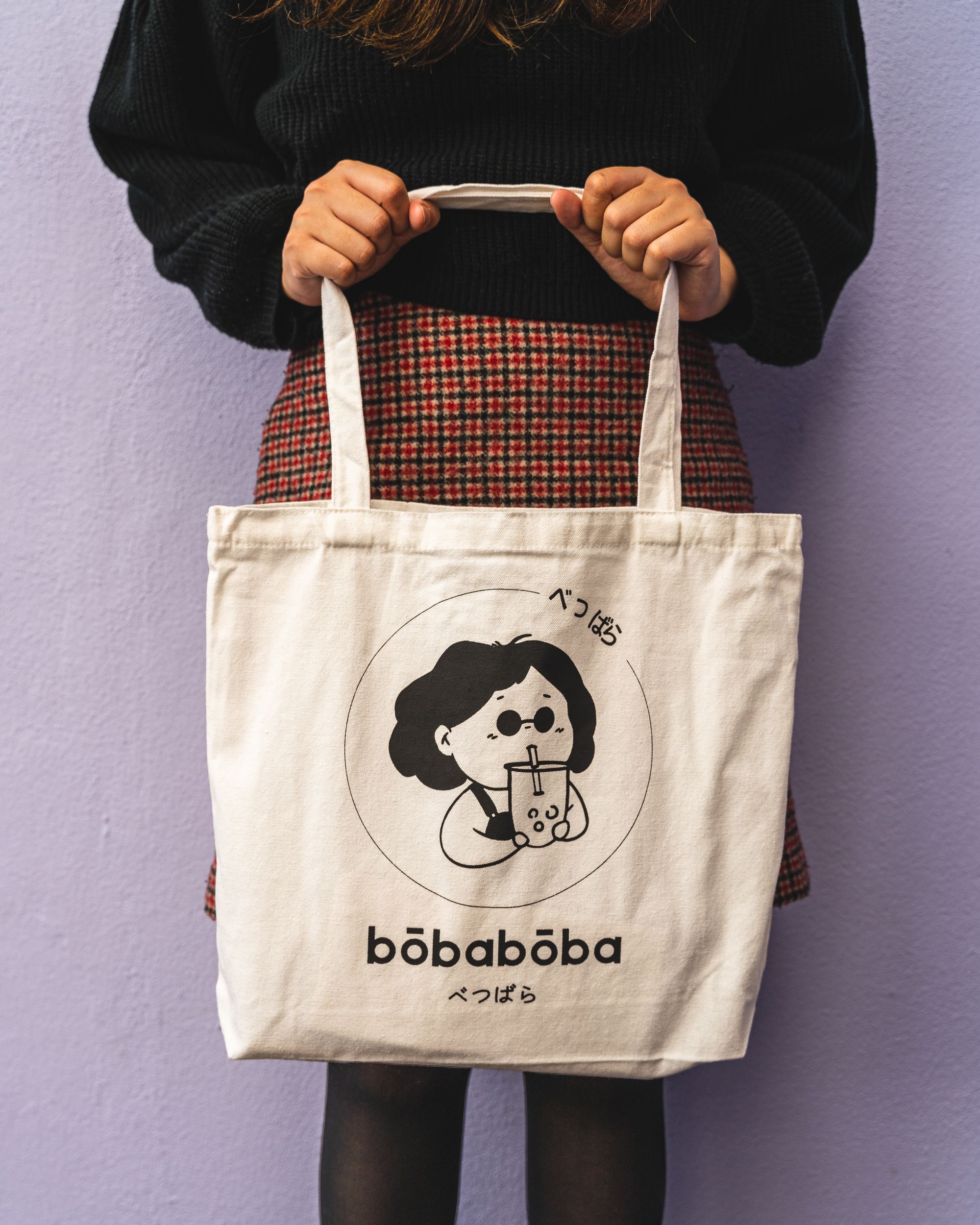 Girl wearing a plaid skirt, tights and a black top holding a white tote bag with a cartoon character sipping bubble tea