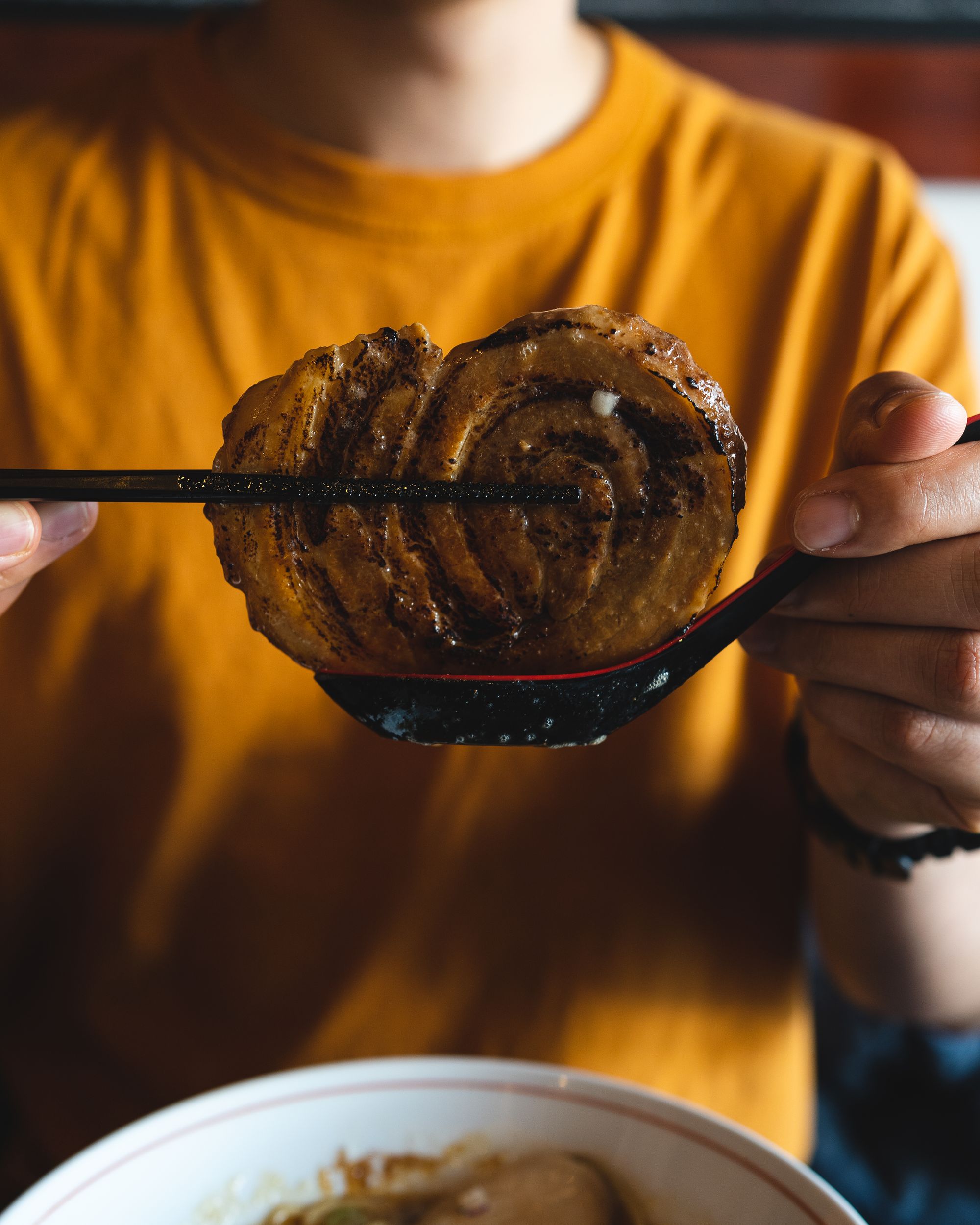 Chopsticks holding up a thick piece of chashu, resting on a soup spoon