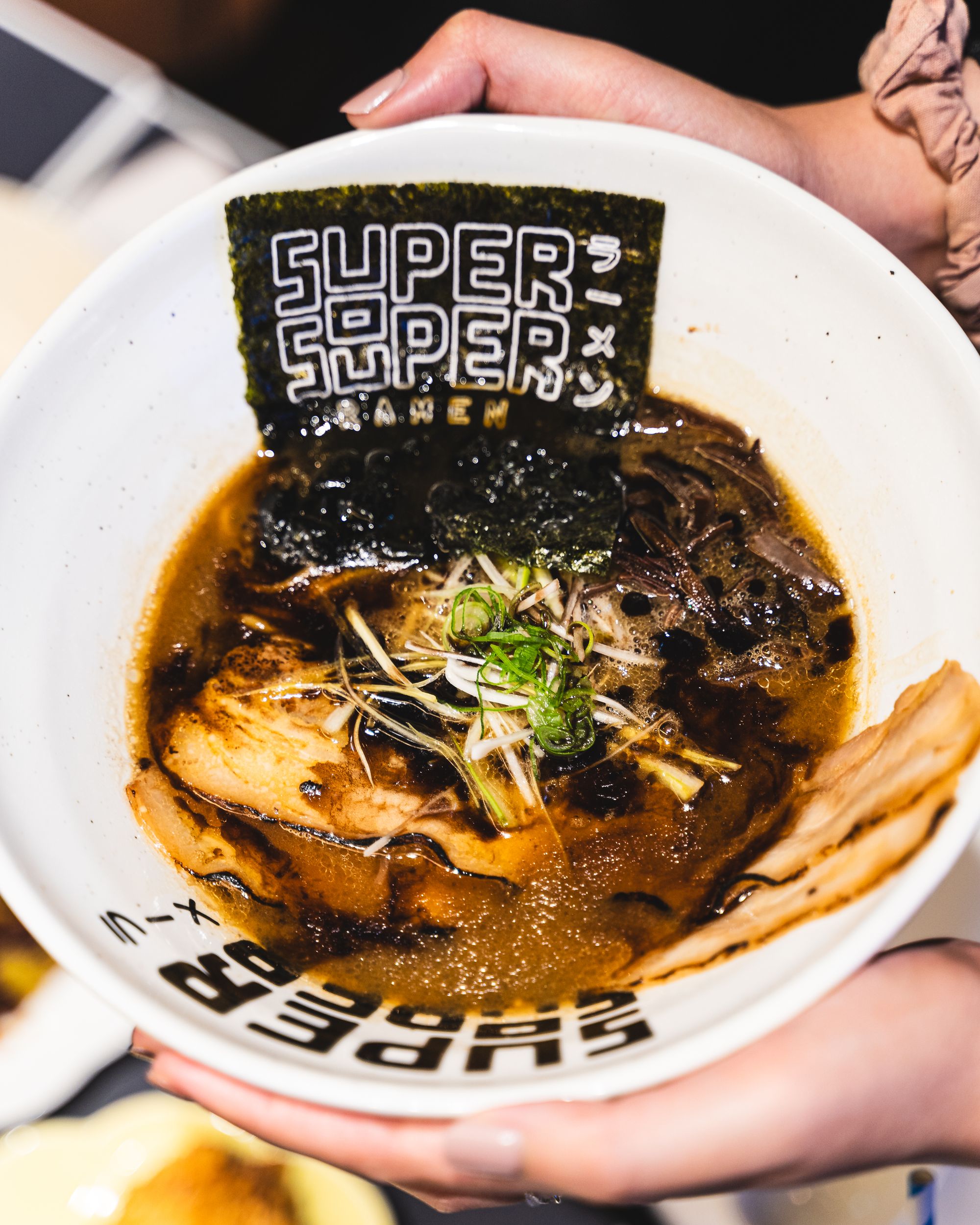 Close up shot of hand holding a bowl of black garlic shoyu ramen, with chashu slices, nori and nori garnish