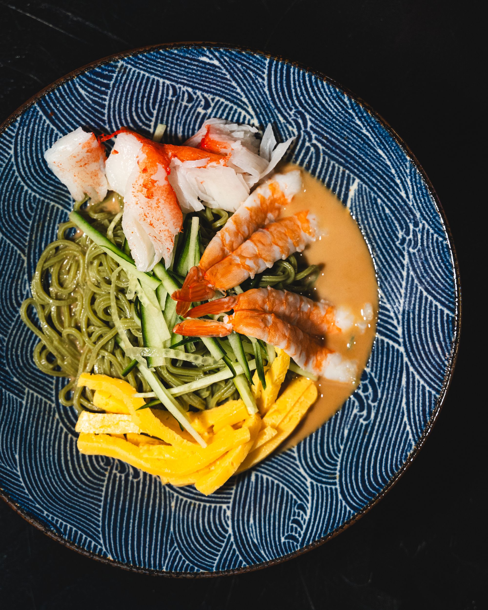 Top down shot of prawn, thinly sliced onion, sliced tamago and crab meat ontop of noodles