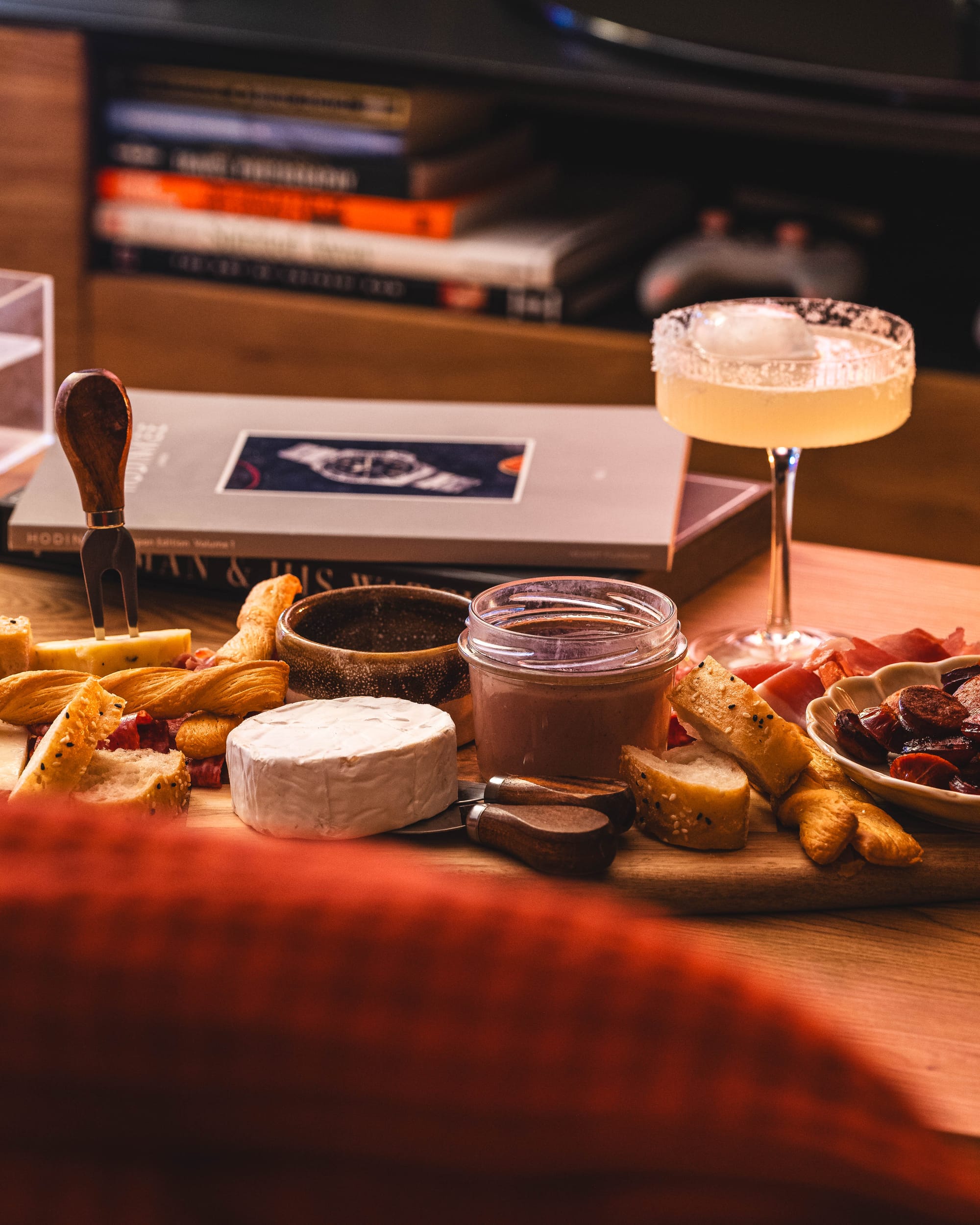 Close up of charcuterie board with brie, bread sticks, cheese, pate in a glass container and a Margherita cocktail in the background