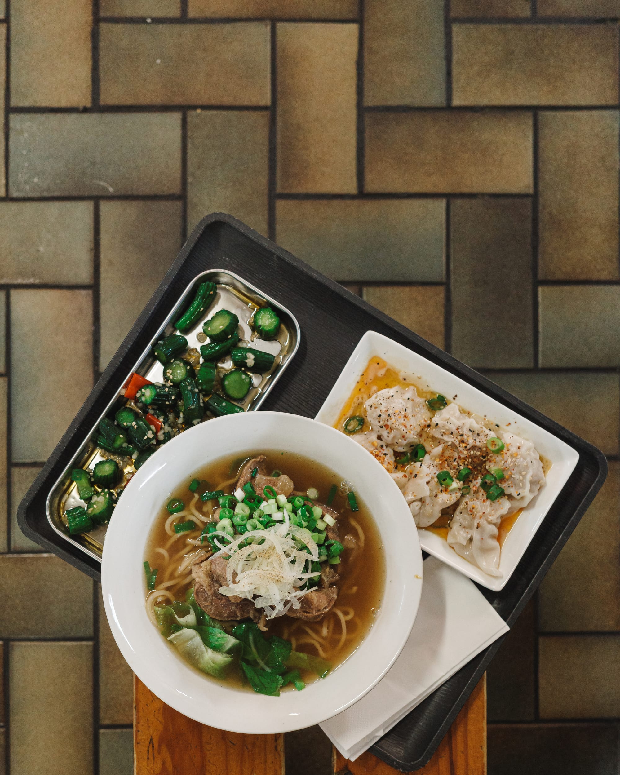 Top down shot of a bowl of beef noodles, plate of wontons and plate of chopped cucumber on a tray