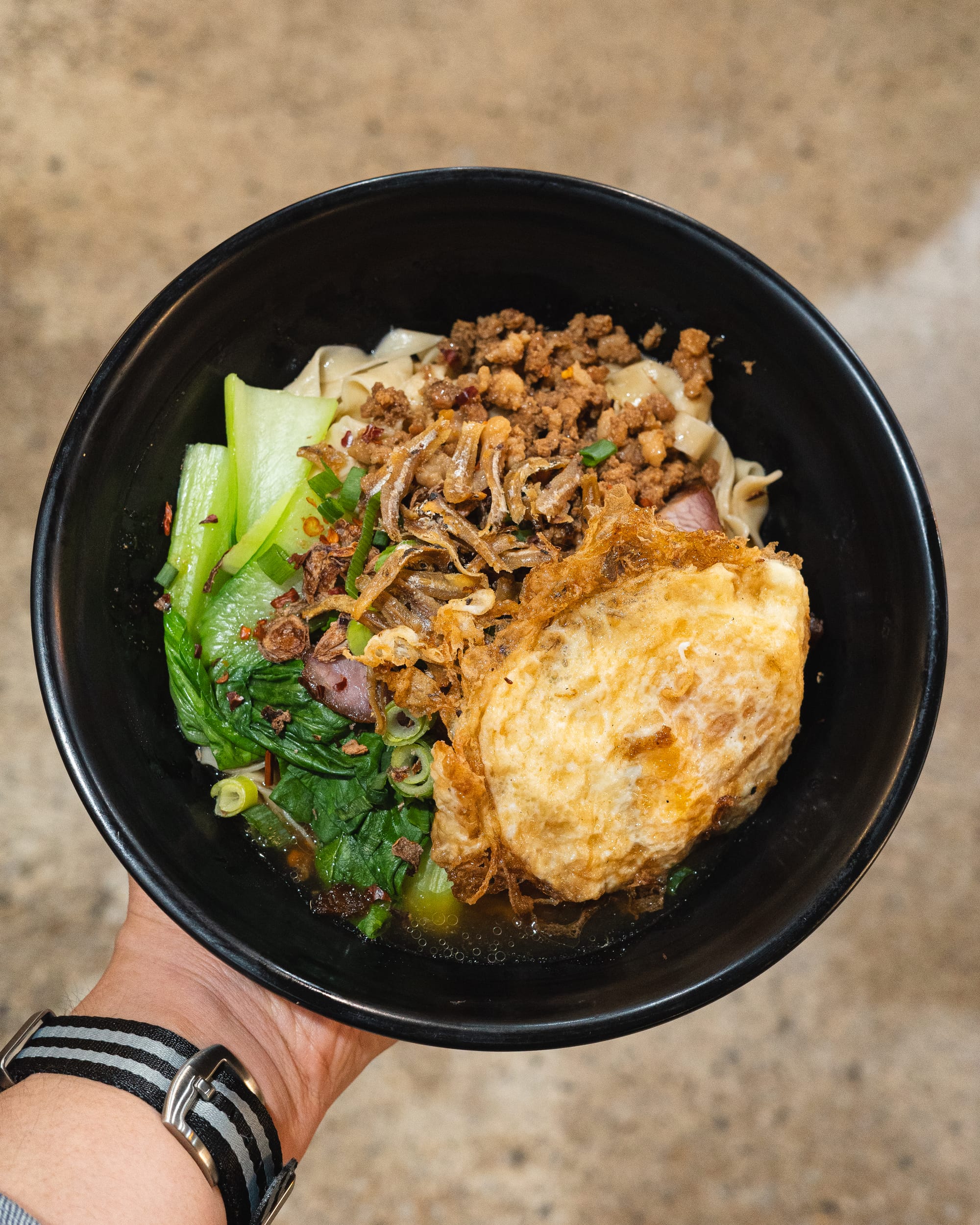 Hand holding a bowl with noodles, BBQ pork, fried egg, fried anchovies and bok choy