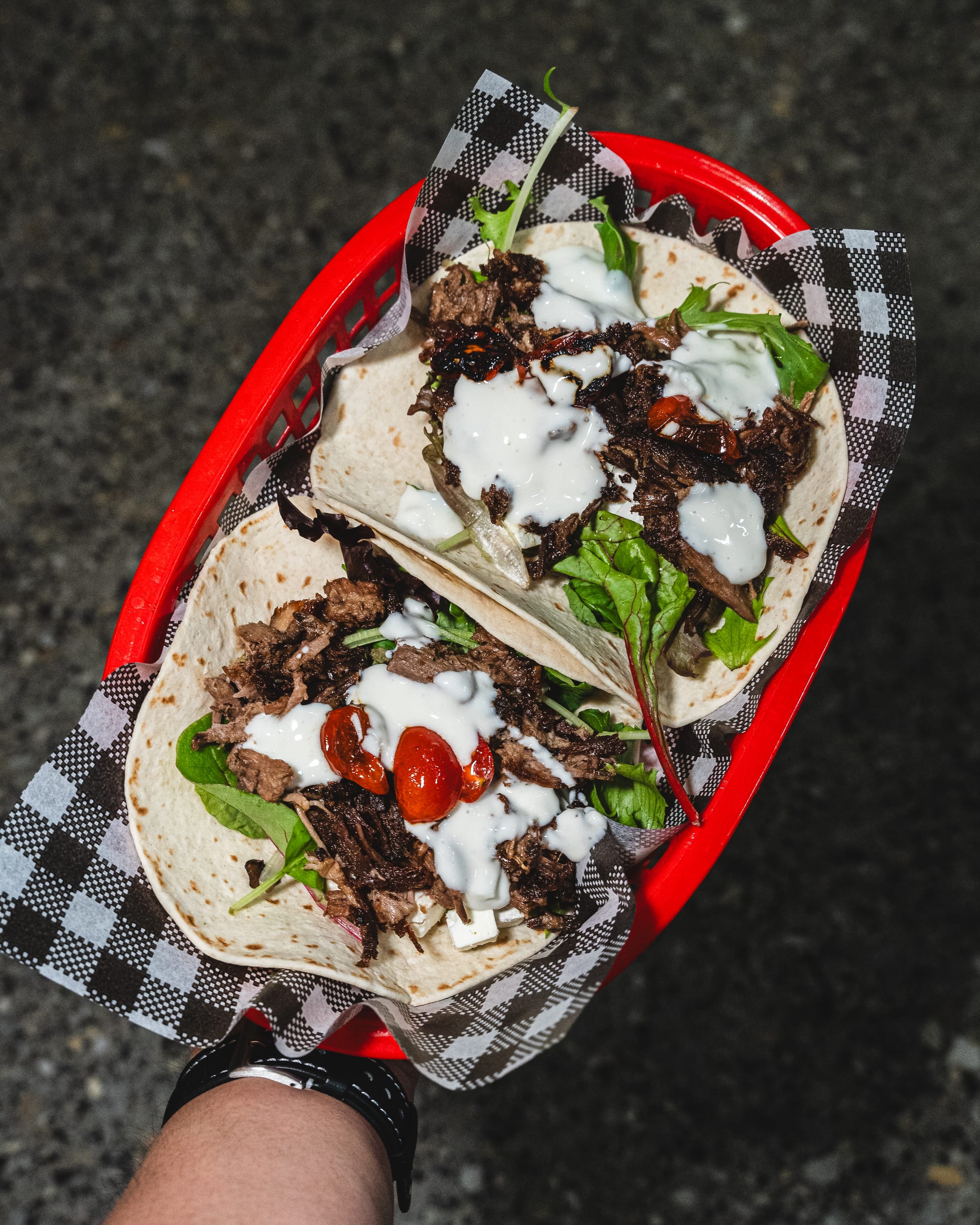Hand holding a tray of tacos with lamb, tzatziki sauce and cherry tomatoes showing