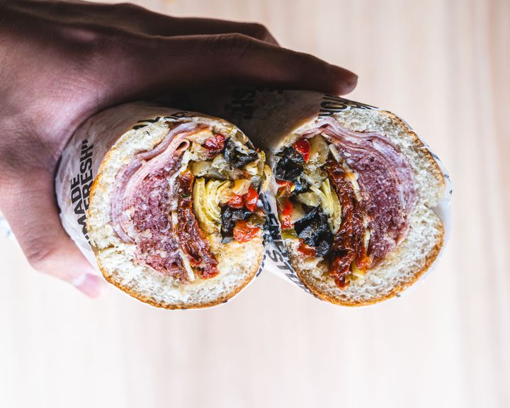 Hand holding two sandwich rolls showing a cross section of Italian cured meats and antipasto