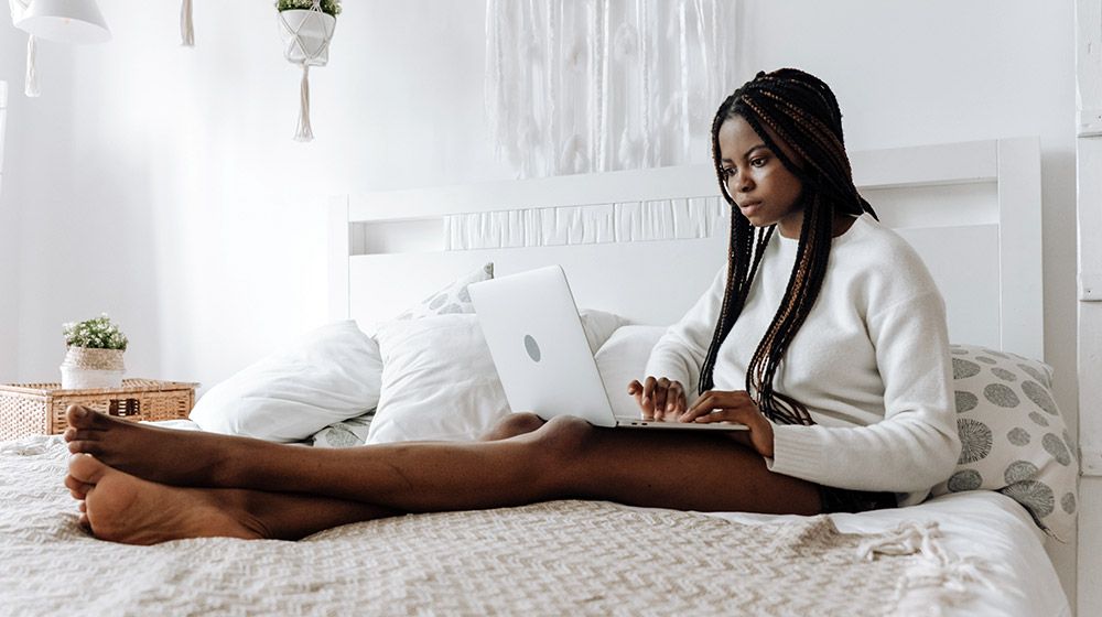 A woman sits on a bed with a laptop in a warm and cozy bedroom