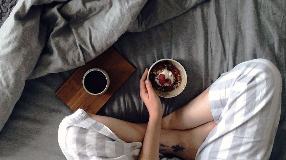 woman in striped pajamas on bed with breakfast bowl and coffee