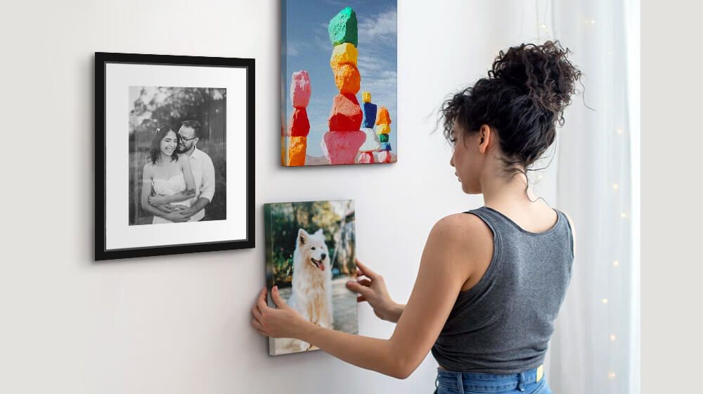 A young woman hangs a 8x10 canvas print on the wall featuring other canvas and framed prints.