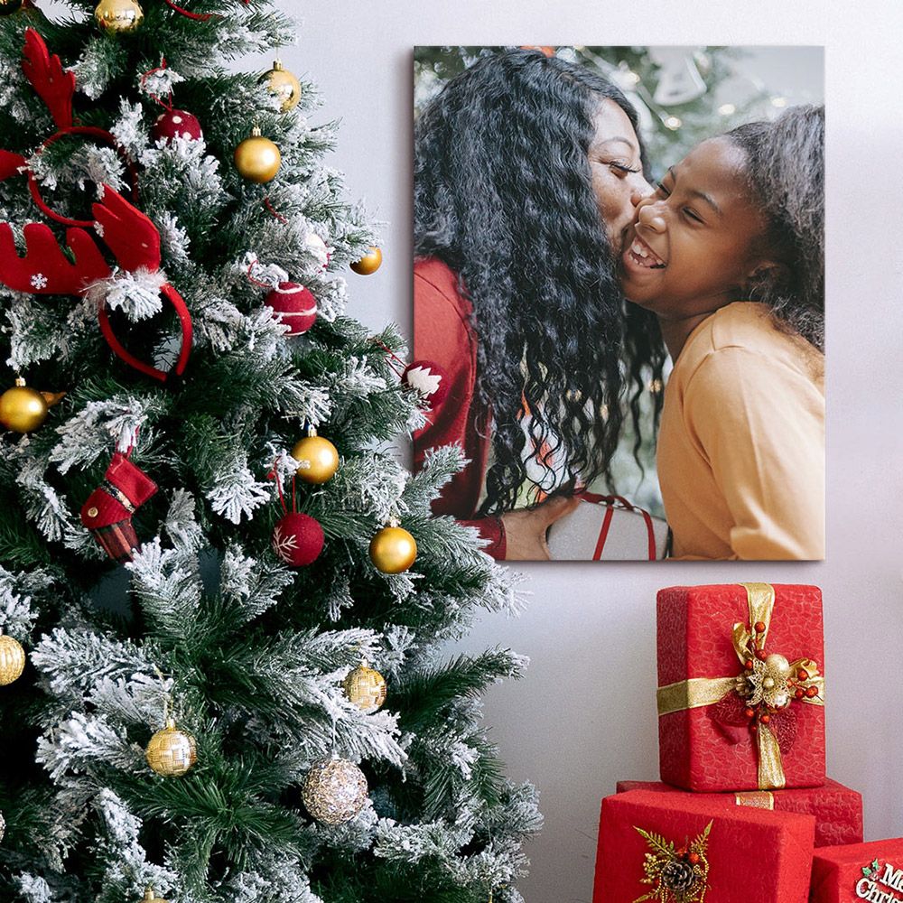 A canvas next to a holiday tree of a woman receiving a gift and kissing a smiling little girl