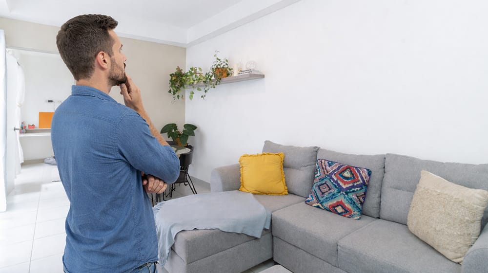 Man standing in living room looking at empty wall above sofa, planning wall decor