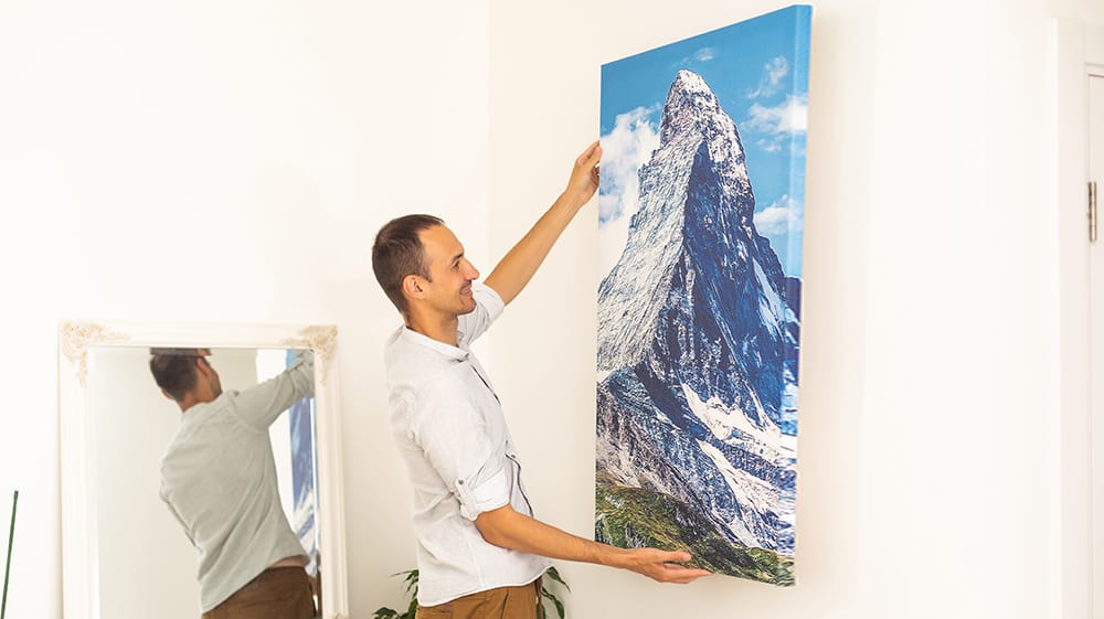 Man hanging a large canvas print of a mountain landscape on a wall