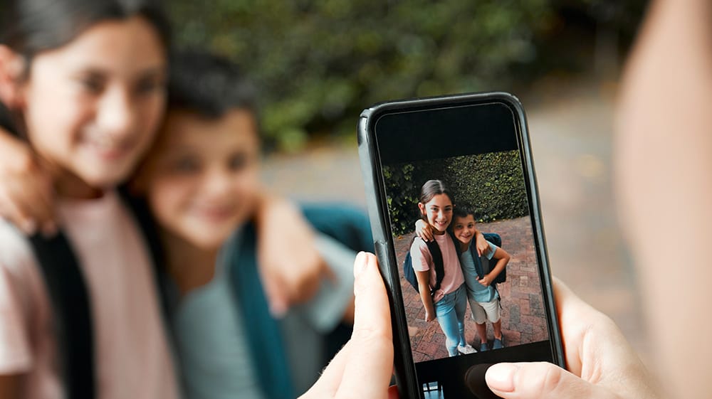 A parent takes a phone photo of two children wearing backpacks and posing together before school.