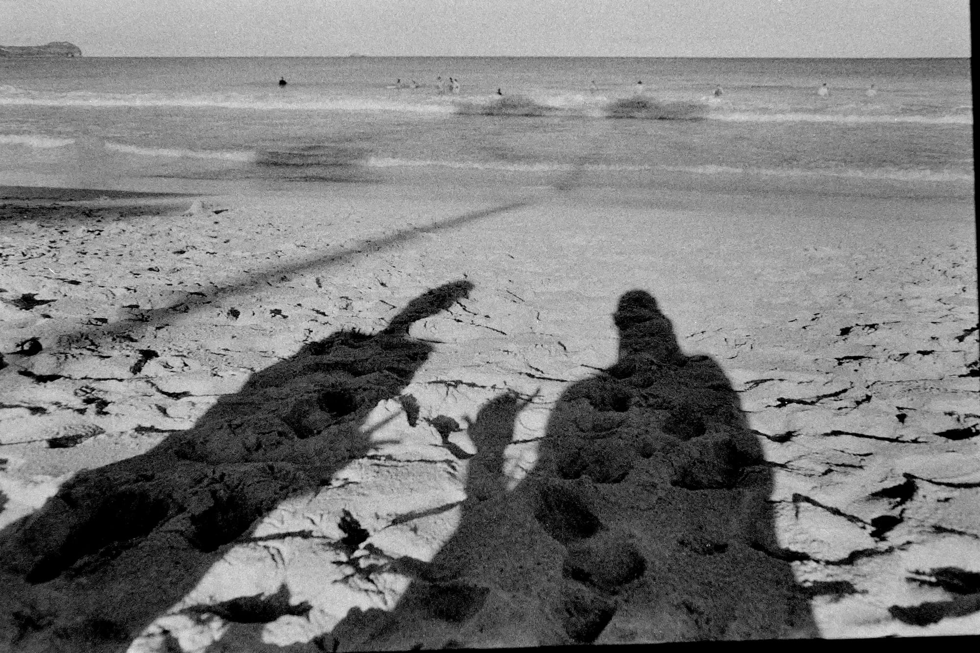 Black and white image of 2 people's shadows on bright sand with the ocean in the background.