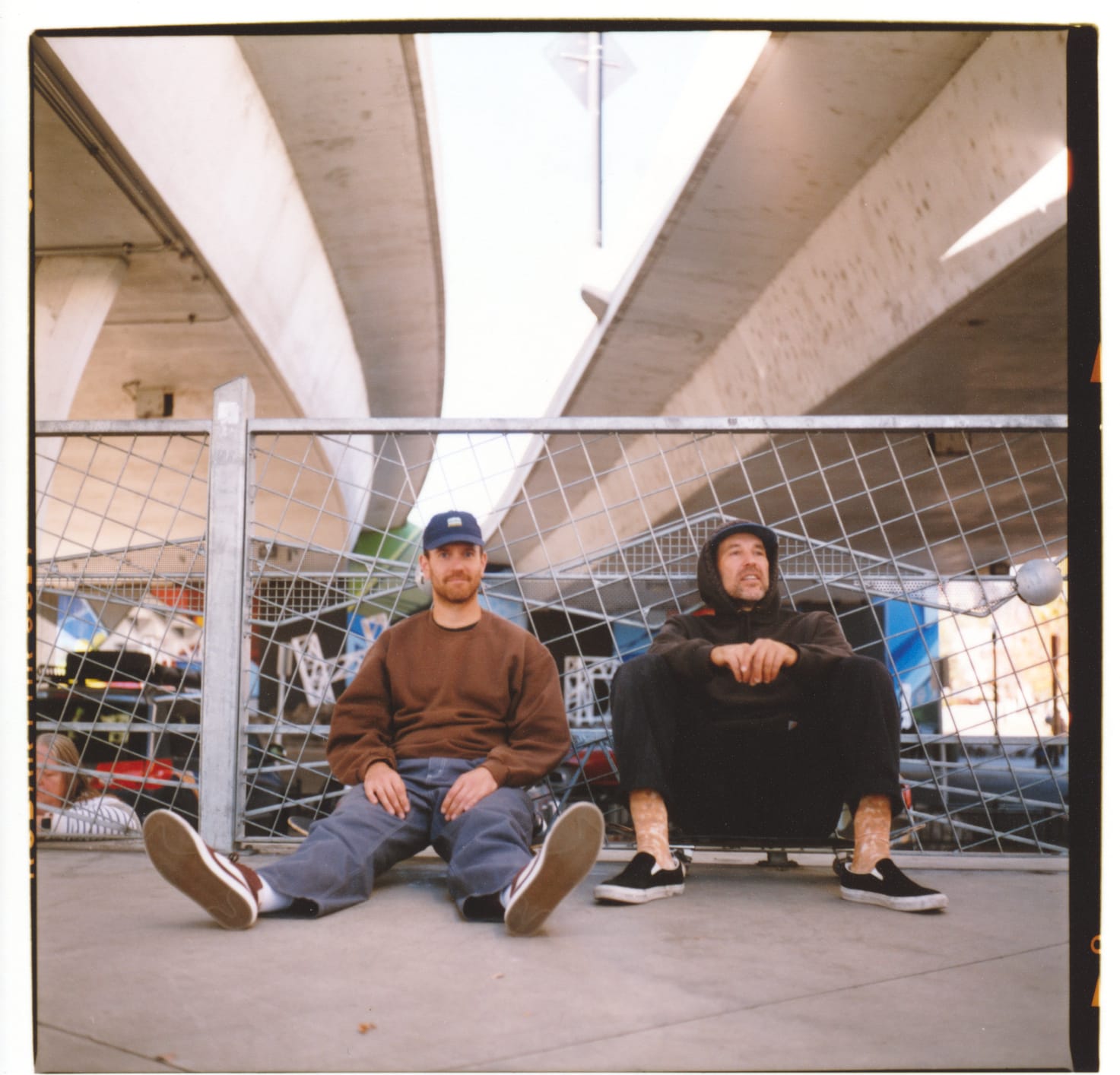 2 guys sitting on their skateboards on the ground with their backs against a fence.
