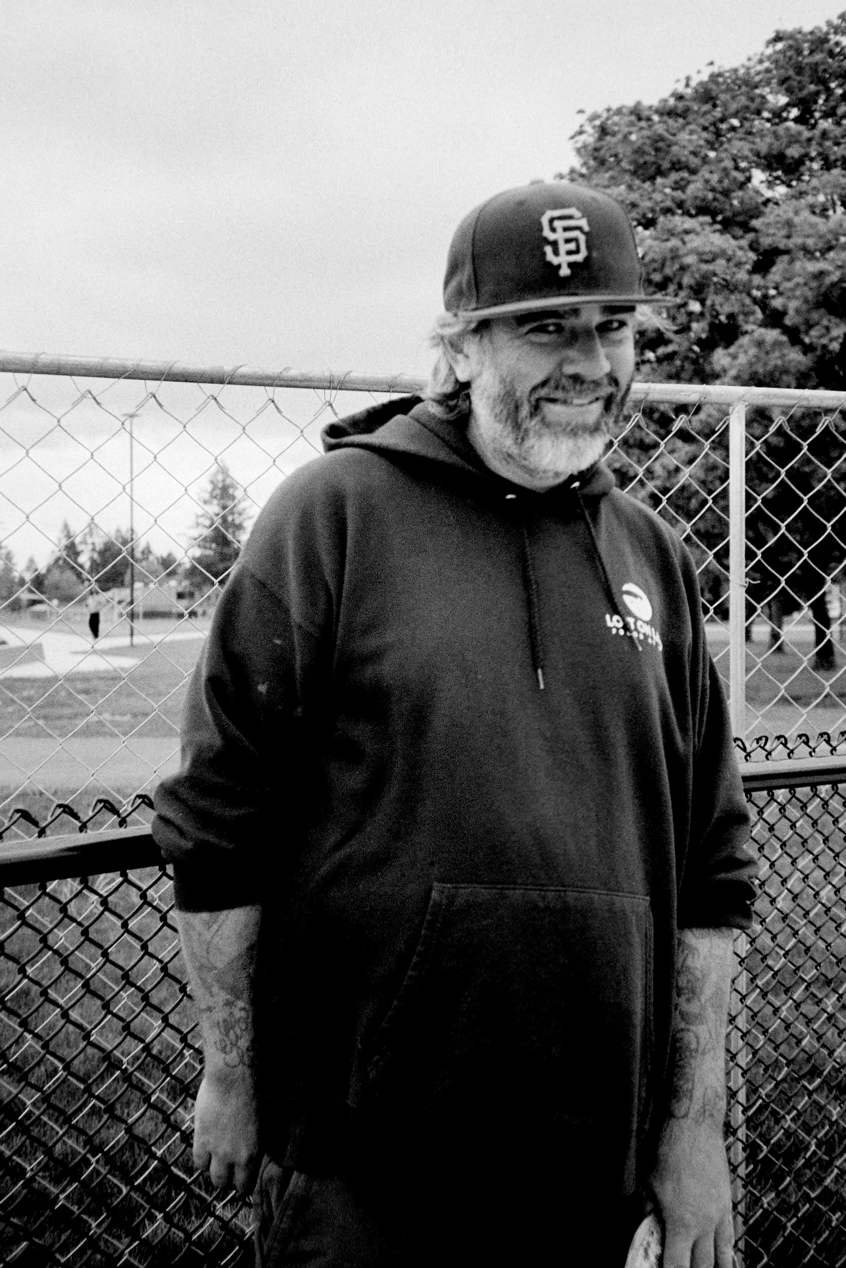 A guy with in a black hoodie and SF hat standing against 2 chainlink fences.