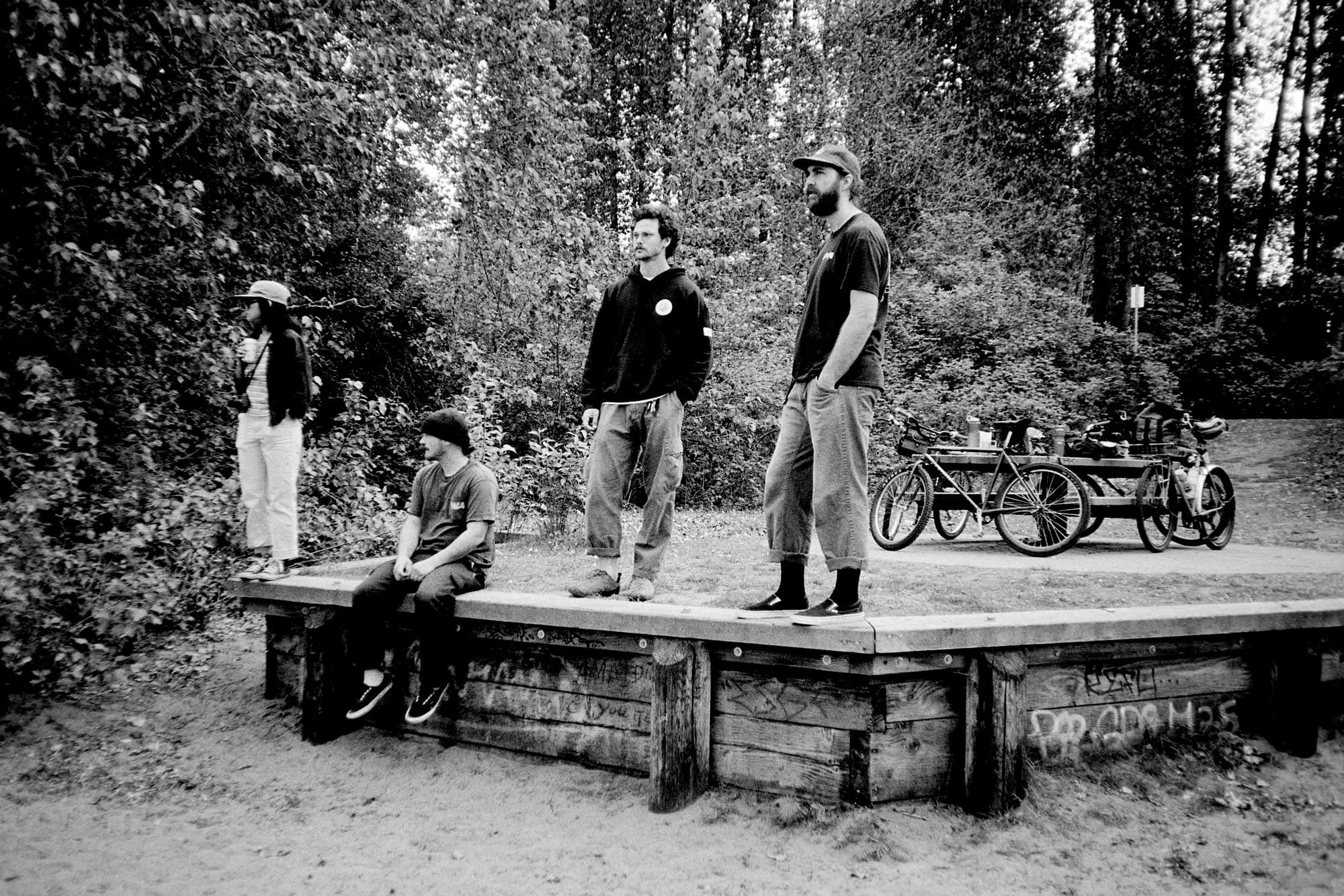 1 woman and 2 men looking to the left. In the background are their bikes leaned against a picnic table with a background of dense trees.