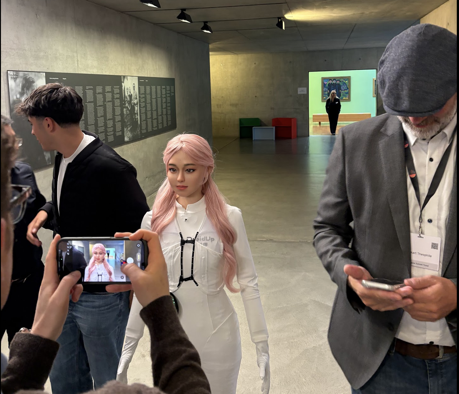 Medium shot of a hyper-realistic humanoid robot with long pink hair and a sleek white suit walking through a minimalist concrete industrial hall. In the foreground, event attendees look at and take smartphone pictures of the robot.