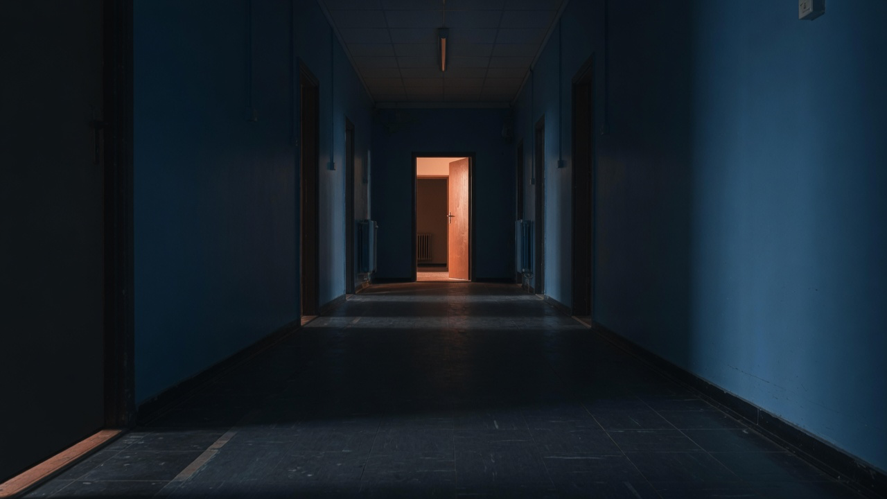 A dark navy corridor receding toward a single warm copper-lit open doorway at the far end, evoking an unfinished but reachable future: the always-on autonomy not yet wired.