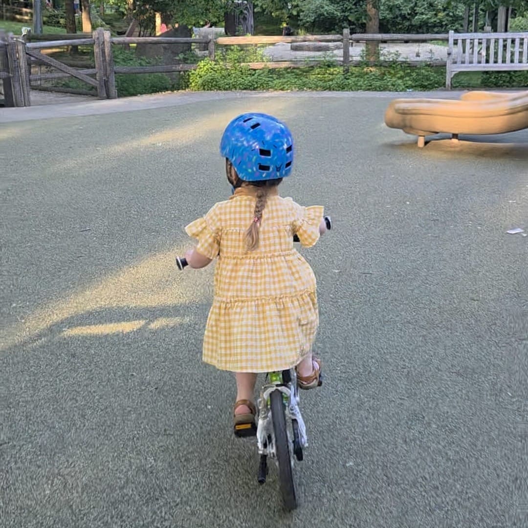 A photograph from behind of a young girl balancing on a bicycle on a sunlit path lined with green trees.