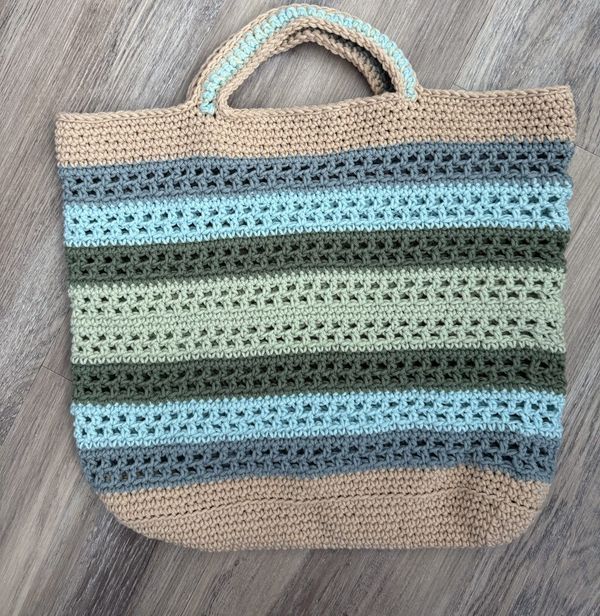 The Coastline Tote, a striped crochet beach bag, on a wood floor