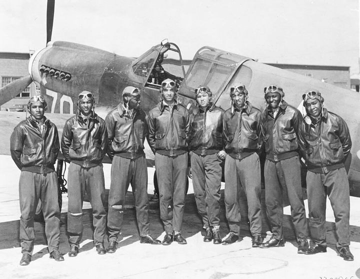 Eight Tuskegee Airmen in flight uniforms and goggles standing in front of a P-40 fighter aircraft