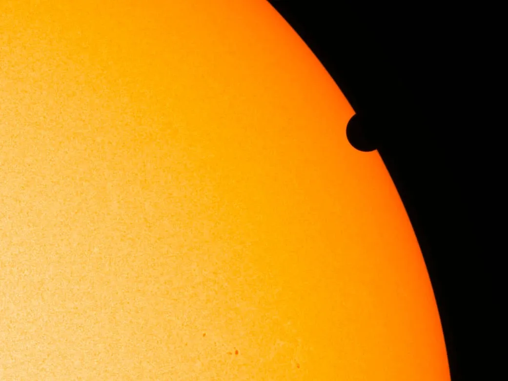 The dark disk of Venus overlaps the right edge of an orange solar disk.