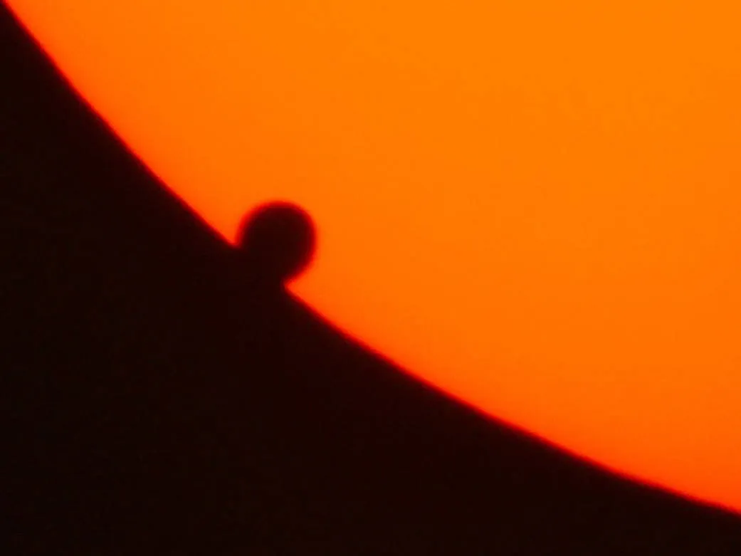 Venus appears as a dark rounded silhouette connected to the edge of an orange solar disk.