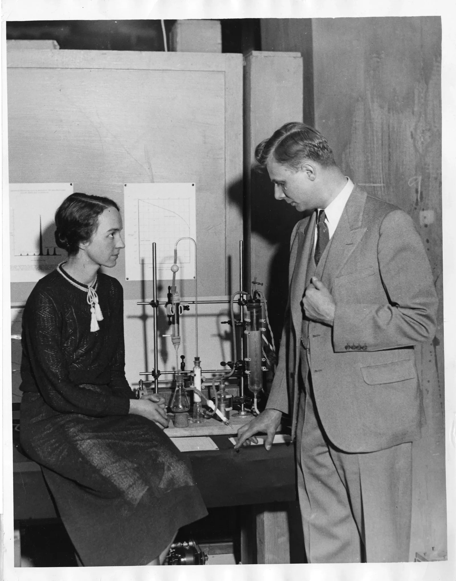 Two physicists stand and sit beside glass apparatus used for making heavy water.