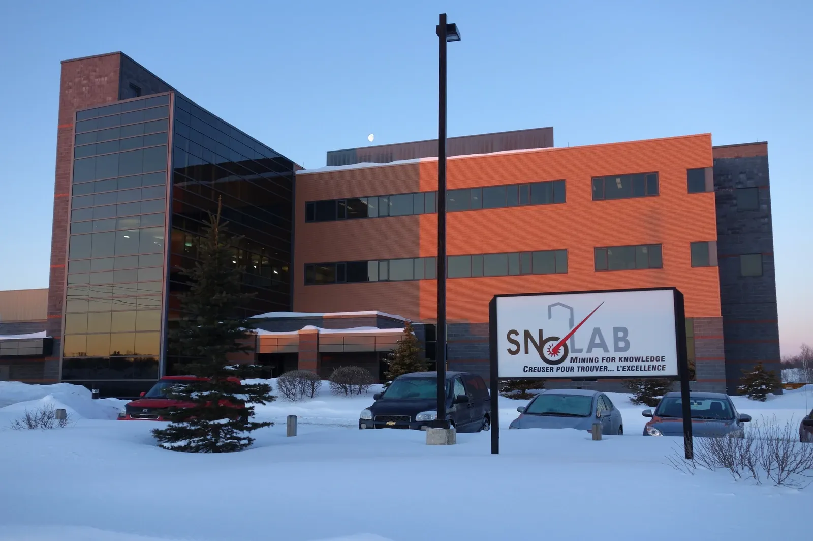 A modern orange and glass laboratory building stands behind snow and a SNOLAB sign.