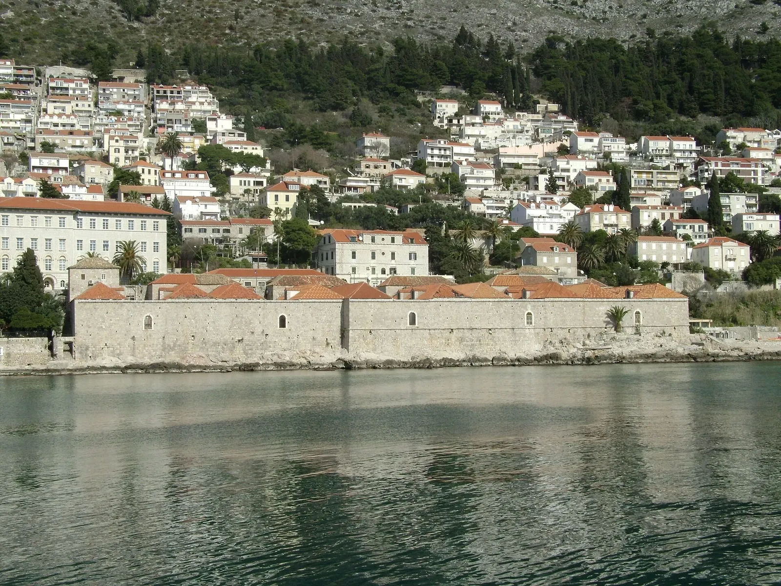 Long stone waterfront buildings of Dubrovnik’s Lazareti with red roofs and the modern hillside city behind them.